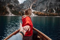 Обратный вид неузнаваемой блондинки, держащейся за руки с кукурузником на деревянной лодке с веслами, плавающими на бирюзовой воде спокойного озера на фоне величественного ландшафта высокогорья в Доломитовых Альпах Италии — стоковое фото