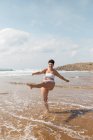 Longitud completa de hembra joven en traje de baño salpicando agua de mar mientras está de pie en la costa arenosa en el día soleado bajo el cielo azul nublado - foto de stock