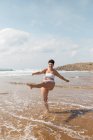 Full length of young female in swimsuit splashing seawater while standing on sandy coast in sunny day under blue cloudy sky — Stock Photo