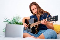 Studentessa concentrata a guardare video tutorial sul laptop mentre impara a suonare la chitarra acustica durante il tempo libero a casa — Foto stock