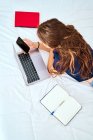 Vue de dessus de dos de l'étudiante méconnaissable couchée sur le lit avec ordinateur portable et manuels scolaires et messagerie sur smartphone pendant les études en ligne à distance à la maison — Photo de stock