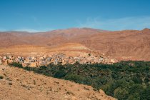 D'en haut de vieux bâtiments en pierre sur la pente de grandes montagnes colorées parmi les plantes vertes avec un ciel bleu clair sur le fond au Maroc — Photo de stock