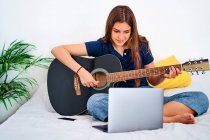 Фокусована студентка-жінка дивиться відеоурок на ноутбуці під час навчання грати на акустичній гітарі під час вільного часу вдома — стокове фото