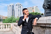 Jovem confiante em terno formal elegante falando no telefone celular e sorrindo enquanto está perto de escultura na praça urbana — Fotografia de Stock