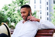 Baixo ângulo de sorrir auto-assegurada jovem barbudo cavalheiro hispânico em roupa elegante e chapéu falando no telefone celular, enquanto descansa no banco na cidade — Fotografia de Stock