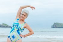Позитивна старша жінка в стильному купальнику робить бічні вправи на вигин, практикуючи здоровий спосіб життя і працюючи на пляжі проти махаючого океану — стокове фото