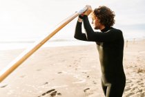 Surfer man dressed in wetsuit standing looking away with the surfboard on the beach during sunrise in the background — Stock Photo