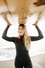 Жінка-серфер, одягнена в гідрокостюм, стоїть, несучи дошку для серфінгу над головою на пляжі під час сходу сонця на задньому плані — стокове фото