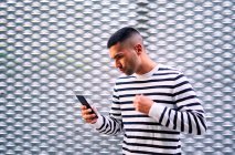 Ein erwachsener hispanischer Mann in Freizeitkleidung steht in der Nähe einer grauen Zierwand und surft auf einem Smartphone in den sozialen Medien auf der Straße der Stadt — Stockfoto