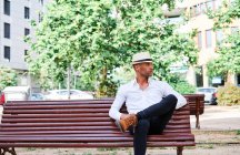 Hombre hispano joven guapo y seguro en ropa elegante y sombrero sentado en el banco y mirando hacia otro lado mientras descansa en la calle de la ciudad - foto de stock