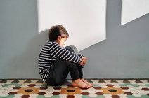 Seitenansicht eines unglücklichen Kindes, das auf dem Boden sitzt und den Kopf mit den Händen bedeckt, während es zu Hause unter häuslicher Gewalt leidet — Stockfoto