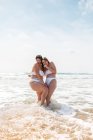 Vista lateral de alegres amigas en trajes de baño abrazándose mientras están de pie en el océano espumoso cerca de la playa de arena bajo el cielo azul nublado en el día soleado - foto de stock