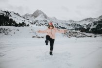 Jovem turista feliz em óculos com perna levantada olhando para a câmera e se divertindo entre o campo na neve perto da montanha — Fotografia de Stock