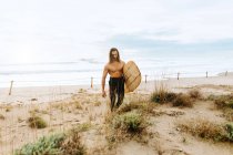 Joven surfista hombre de pelo largo vestido con traje de neopreno caminando mirando hacia otro lado con tabla de surf en dunas arenosas - foto de stock