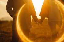 Namoradas sem rosto de mãos dadas suavemente em pé na chama da lente de luz do pôr do sol na natureza — Fotografia de Stock