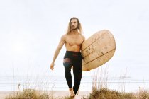 Homem surfista jovem com cabelo comprido vestido de fato de mergulho a andar com o olhar para a prancha de surf em dunas de areia — Fotografia de Stock