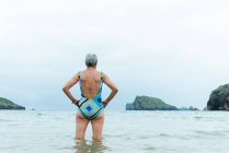 Back view active fit aged female in swimsuit walking out of sea water while enjoying summer day on beach — Stock Photo