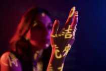 Jeune modèle féminin à la mode avec projection de lumière en forme de hiéroglyphes orientaux regardant main tendue dans un studio sombre avec éclairage rouge — Photo de stock