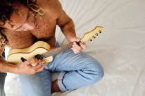 Сверху талантливый музыкант с обнаженным туловищем и вьющимися волосами сидит на кровати и играет на укулеле — стоковое фото