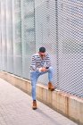 Homem hispânico de corpo inteiro em roupa elegante olhando para longe e usando o celular enquanto se inclina na parede na rua da cidade — Fotografia de Stock