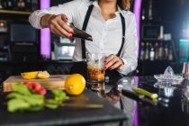 Cropped unrecognizable female barkeeper in stylish outfit adding ice cubes into glass while preparing mojito cocktail standing at counter in modern bar — Stock Photo