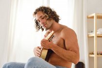 Vista lateral de talentoso músico masculino con el torso desnudo y el pelo rizado sentado en la cama y tocando ukelele - foto de stock