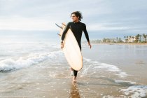 Surfer im Neoprenanzug geht mit Surfbrett in Richtung Wasser, um bei Sonnenaufgang eine Welle am Strand zu fangen — Stockfoto