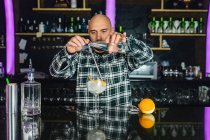 Орієнтований чоловічий бармен додає рідину з пляшки в склянку з довгою ложкою, готуючи коктейль, що стоїть за стійкою в сучасному барі — стокове фото