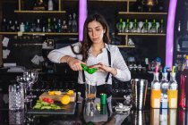 Jeune femme barman en tenue élégante serrant du citron tout en préparant un cocktail mojito debout au comptoir dans un bar moderne — Photo de stock
