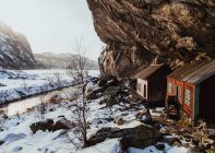 Cabanes vieillies près des parois rocheuses entre les terres sauvages dans la neige près de la rivière étroite et le ciel bleu — Photo de stock