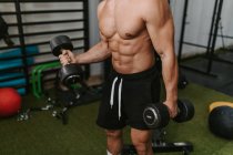 Cropped unrecognizable muscular young male trainer with naked torso lifting heavy dumbbells while training in gym — Stock Photo