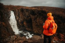 Вид ззаду молодого туриста в зимовому одязі, який дивиться на каскад і гірську річку між кам'яним пагорбом — стокове фото