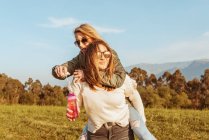 Laughing blond woman giving piggyback ride to cheerful girlfriends holding soap bubble stick walking in nature — Stock Photo