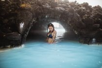 Side view of young tattooed hipster in swimsuit posing in blue water between rocks — Stock Photo