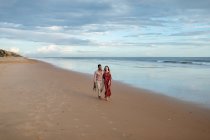 Alegre pareja multirracial cogida de la mano y caminando a lo largo de la orilla húmeda mientras admira el mar al atardecer en verano - foto de stock