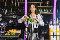 Молодая барменша в стильном наряде, сжимающая лимон, пока готовит коктейль, стоя у стойки в современном баре — стоковое фото