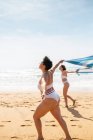 Full body side view of female friends in swimsuits strolling on sandy shore with towel near ocean under blue cloudy sky in sunny day — Stock Photo