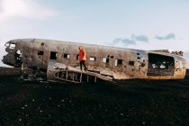 Side view of young tourist standing on wrecked aircraft between deserted lands and blue sky — Stock Photo