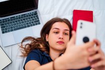 Зверху позитивна молода студентка лежить на ліжку і приймає селфі на смартфон, маючи перерву під час дистанційного онлайн-навчання вдома — стокове фото