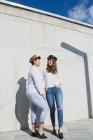 Cuerpo completo de jóvenes amigas positivas en trajes de moda y sombreros de pie mirándose unas a otras en la pasarela cerca de la pared gris en un día soleado bajo el cielo azul - foto de stock
