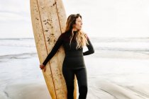 Surfista mulher vestida de fato de mergulho de pé olhando para longe com a prancha de surf na praia durante o nascer do sol no fundo — Fotografia de Stock