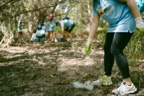 Анонимный активист в защитных перчатках собирает пластиковую бутылку с земли, собирая мусор в природе — стоковое фото