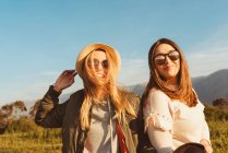 Young close female friends in stylish clothes standing together on meadow in mountains looking at camera in golden light — Stock Photo