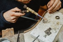 Anonymous goldsmith using blowtorch to heat tiny metal ornament while making jewellery on workbench — Stock Photo