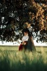 Jovem fêmea em blusa branca à moda antiga e saia segurando cesta de vime cheia de maçãs frescas enquanto descansa perto da árvore no dia de verão no campo — Fotografia de Stock