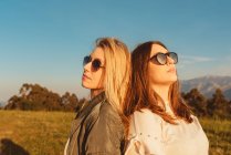 Young close female friends in stylish clothes standing together on meadow in mountains looking away in golden light — Stock Photo