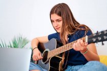 Фокусована студентка-жінка дивиться відеоурок на ноутбуці під час навчання грати на акустичній гітарі під час вільного часу вдома — стокове фото