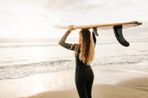 Вид ззаду на невизначений жіночий серфер, одягнений у гідрокостюм, що стоїть, тримаючи дошку для серфінгу на голові на пляжі під час сходу сонця на задньому плані — стокове фото