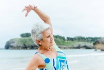 Mujer senior positiva en traje de baño elegante haciendo ejercicio de curva lateral mientras practica un estilo de vida saludable y hace ejercicio en la playa contra el océano ondulante - foto de stock