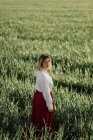 Calm young female dressed in old fashioned blouse and skirt standing alone among tall green grass in cloudy summer day in countryside — Stock Photo